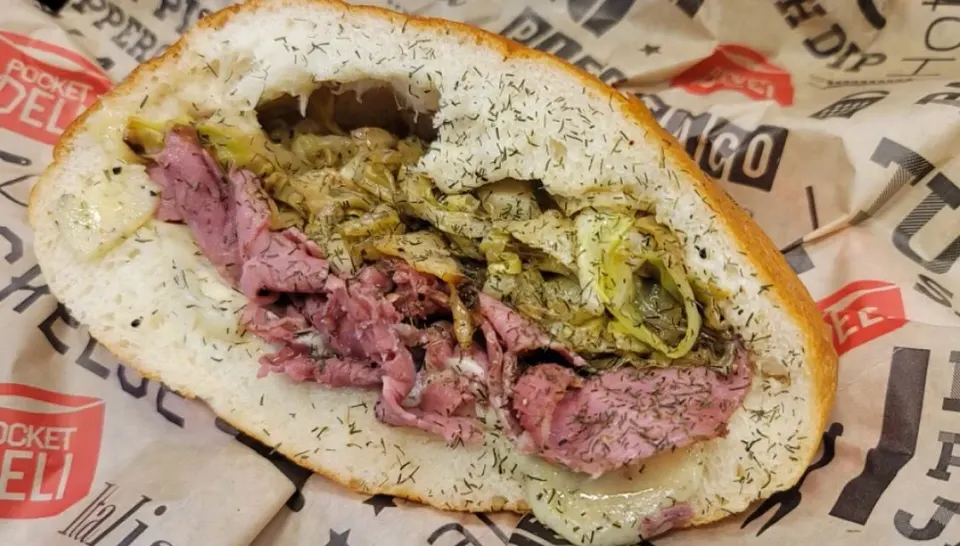 Italian beef sandwich with sliced beef, pickles, and herbs on a hoagie roll wrapped in branded paper.